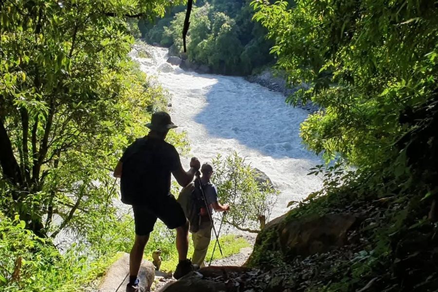 Trekkers walking along trail beside rushing Langtang Khola River surrounded by lush green forest